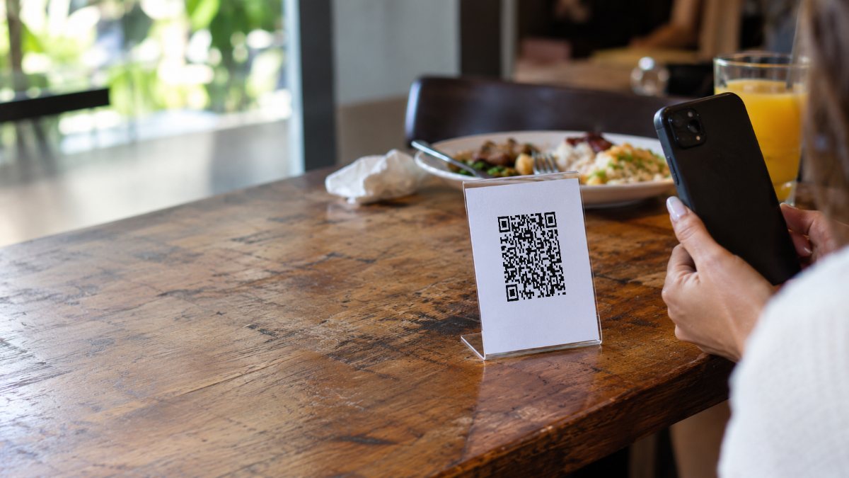 Código QR en un soporte de acrílico sobre la mesa de un restaurante, y un comensal apuntándole con el teléfono.